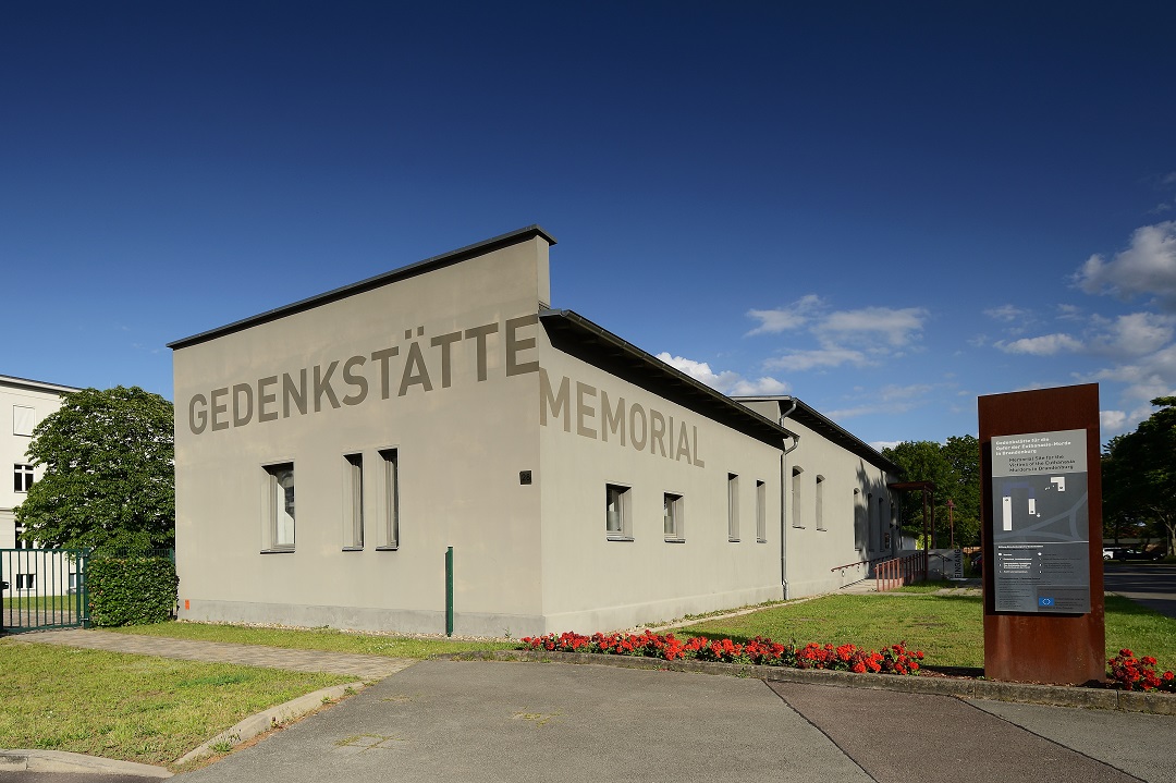 Ein graues einstöckiges Gebäude. Darauf die Aufschrift "Gedenkstätte / Memorial". Rechts im Bild eine rostige Stele mit Informationen.