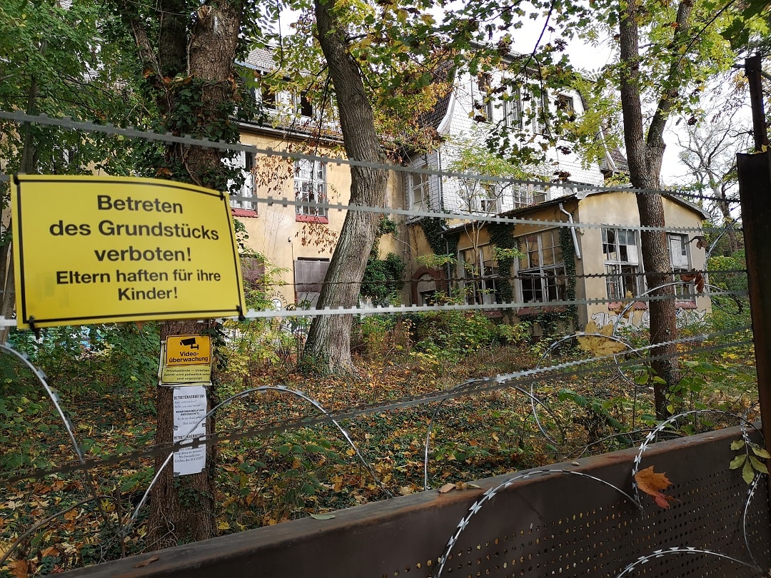 Ein Zaun mit Stacheldraht. Dahinter ein Gebäude. Am Zaun ein Schild mit der Aufschrift "Betreten des Grundstücks verboten! Eltern haften für ihre Kinder!" thumbnail