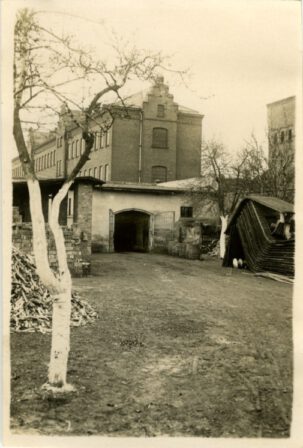 Im Vordergrund eine Scheune mit geöffnetem Tor. Im Hintergrund und am rechten Bildrand Zellengebäude.