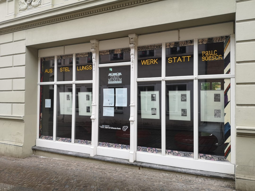 Ein altes, mehrgliedriges Schaufenster in einem Altbau. Umlaufend ein aufgeklebter Rahmen aus Bildelementen Paul Goeschs. Oben die Überschrift: Ausstellungswerkstatt Paul Goesch. In jedem der Fensterteile hängt ein Bilderrahmen, darin Druckgrafiken.