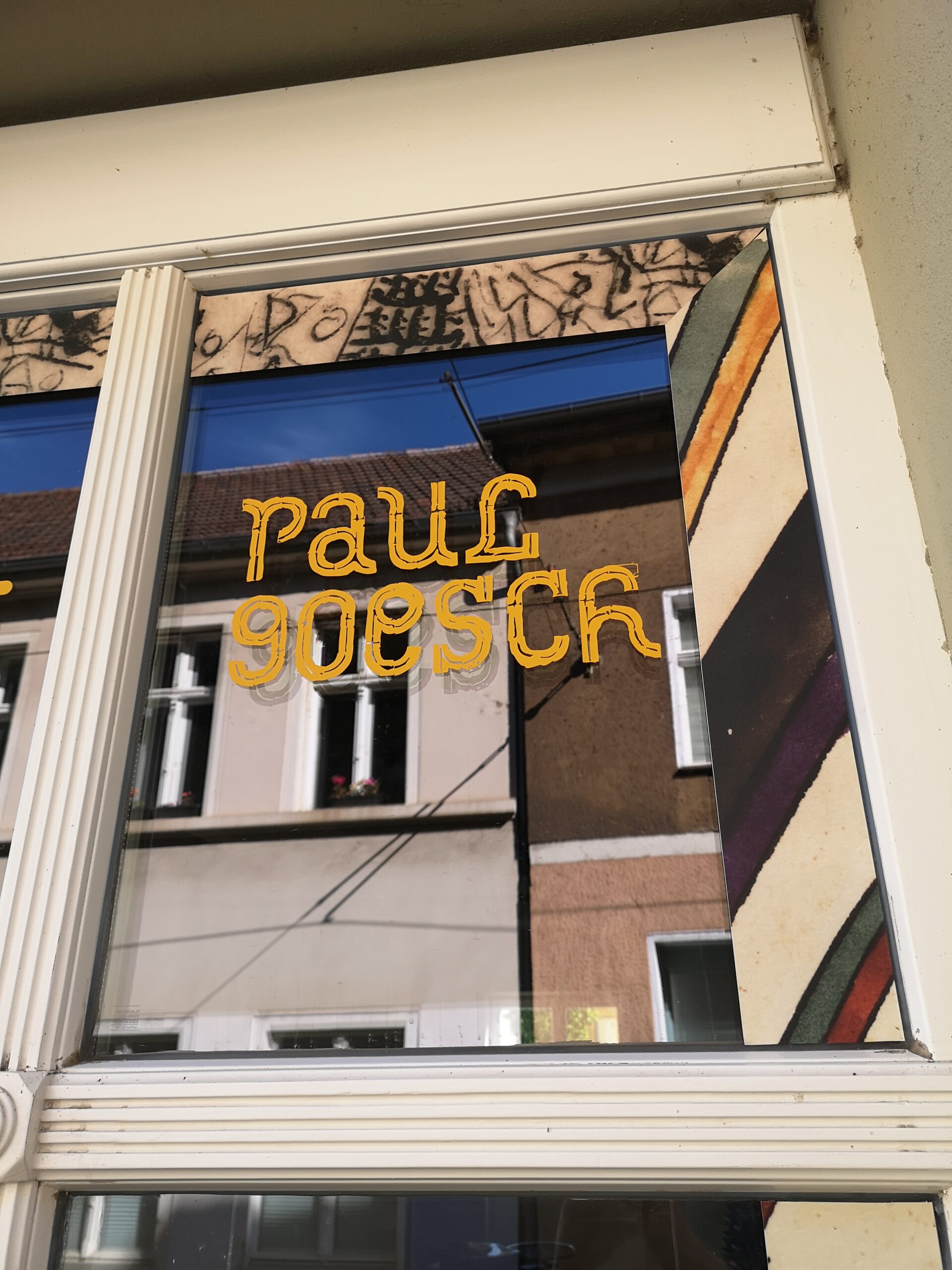 Ausschnitt eines alten Fensters. Auf die Scheibe ist umlaufend ein Rahmen aus Bildelement aus Goeschs Werken aufgeklebt. In der Mitte steht in gelber, verschnörkelter Schrift: "Paul Goesch"
