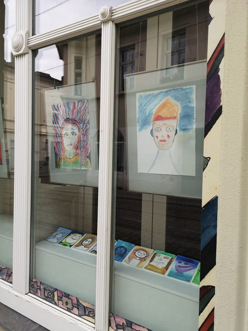 Ausschnitt des Schaufensters. Hinter der Scheibe sind in Rahmen gehängte, großformatige Aquarellporträts zu sehen