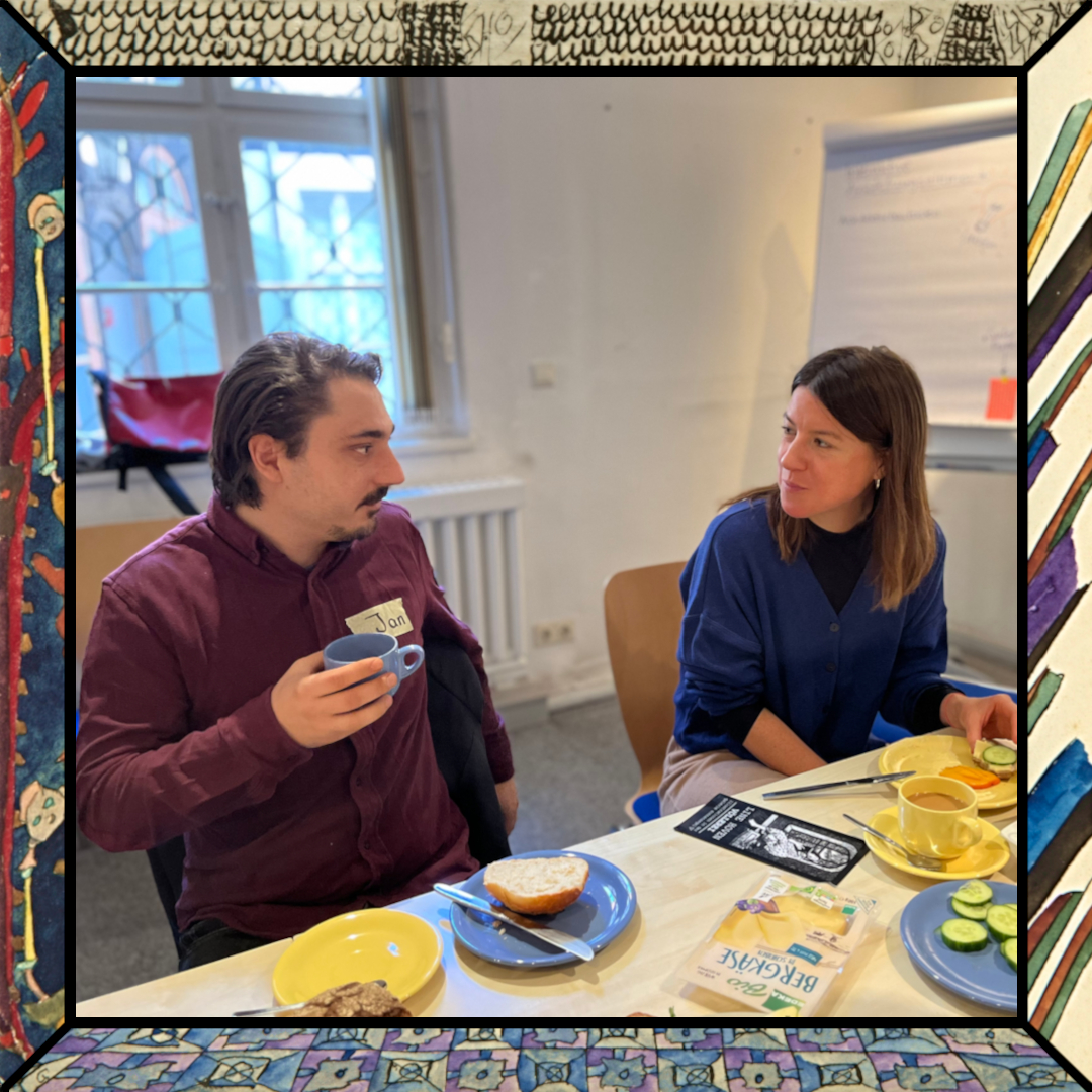 Ein Mann und eine Frau an einem gedeckten Frühstckstisch, in ein Gespräch vertieft. Er hält eine Kaffeetasse