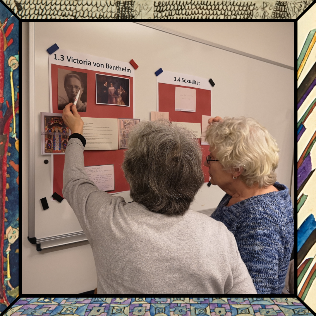 Zwei ältere Frauen zeigen auf ein rotes Plakat, dem Betrachter den Rücken zugewandt