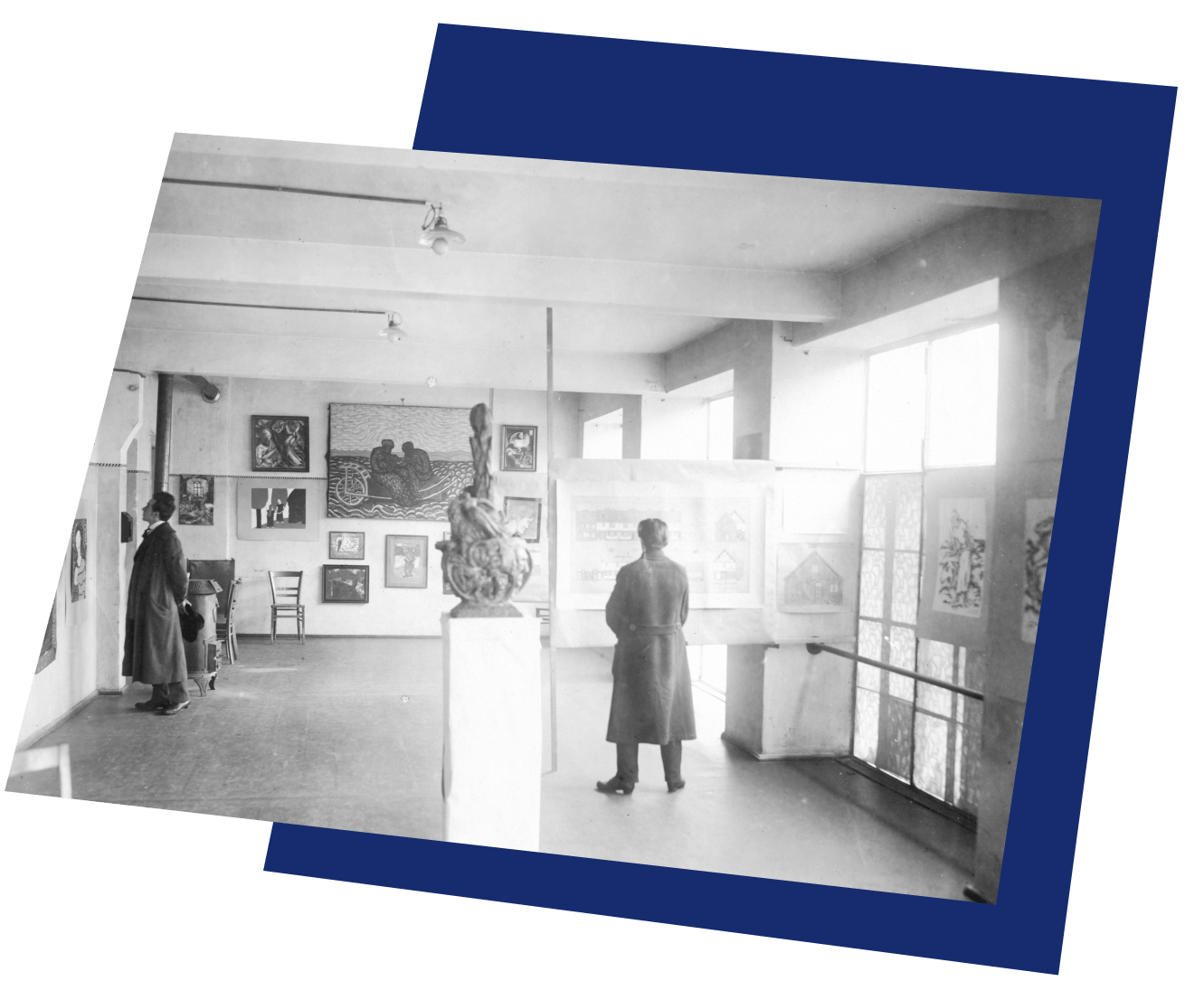 Ein altes Foto: ein Ausstellungsraum voller Bilder und einer Installation. 2 Personen stehen im Raum und schauen sich Bilder an. Die beiden Personen haben beide lange Jacken an und stehen weit voneinander entfernt.