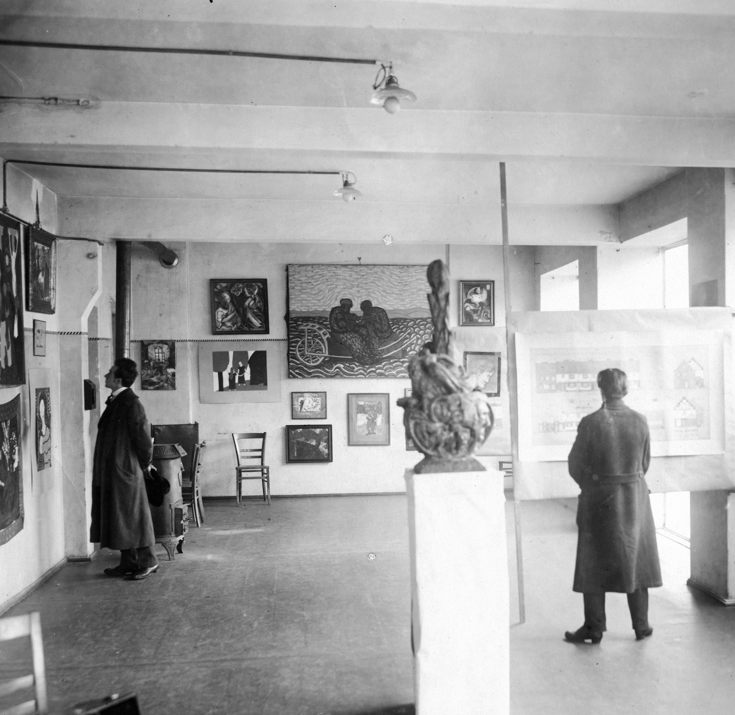 Ein altes Foto: ein Ausstellungsraum voller Bilder und einer Installation. 2 Personen stehen im Raum und schauen sich Bilder an. Die beiden Personen haben beide lange Jacken an und stehen weit voneinander entfernt.