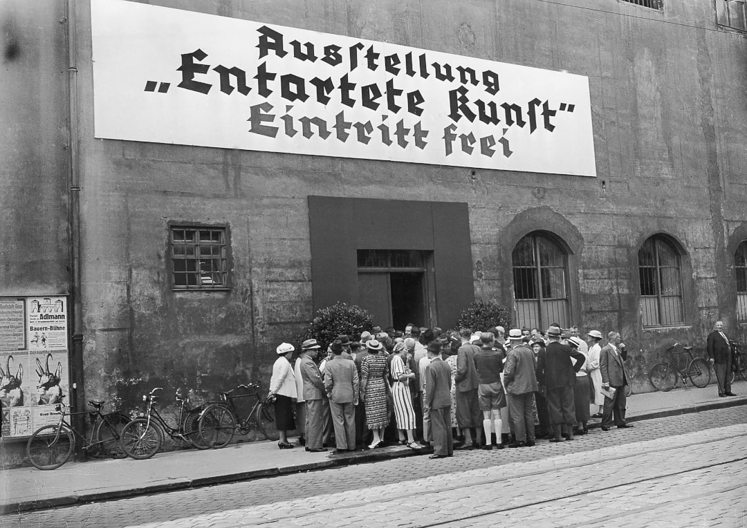 Ein altes Foto. Vor einem großen Haus steht eine Gruppe von ca. 50 Menschen. An dem Haus hängt eine goße Werbetafel, auf der steht: Ausstellung "Entartete Kunst" Eintritt frei.